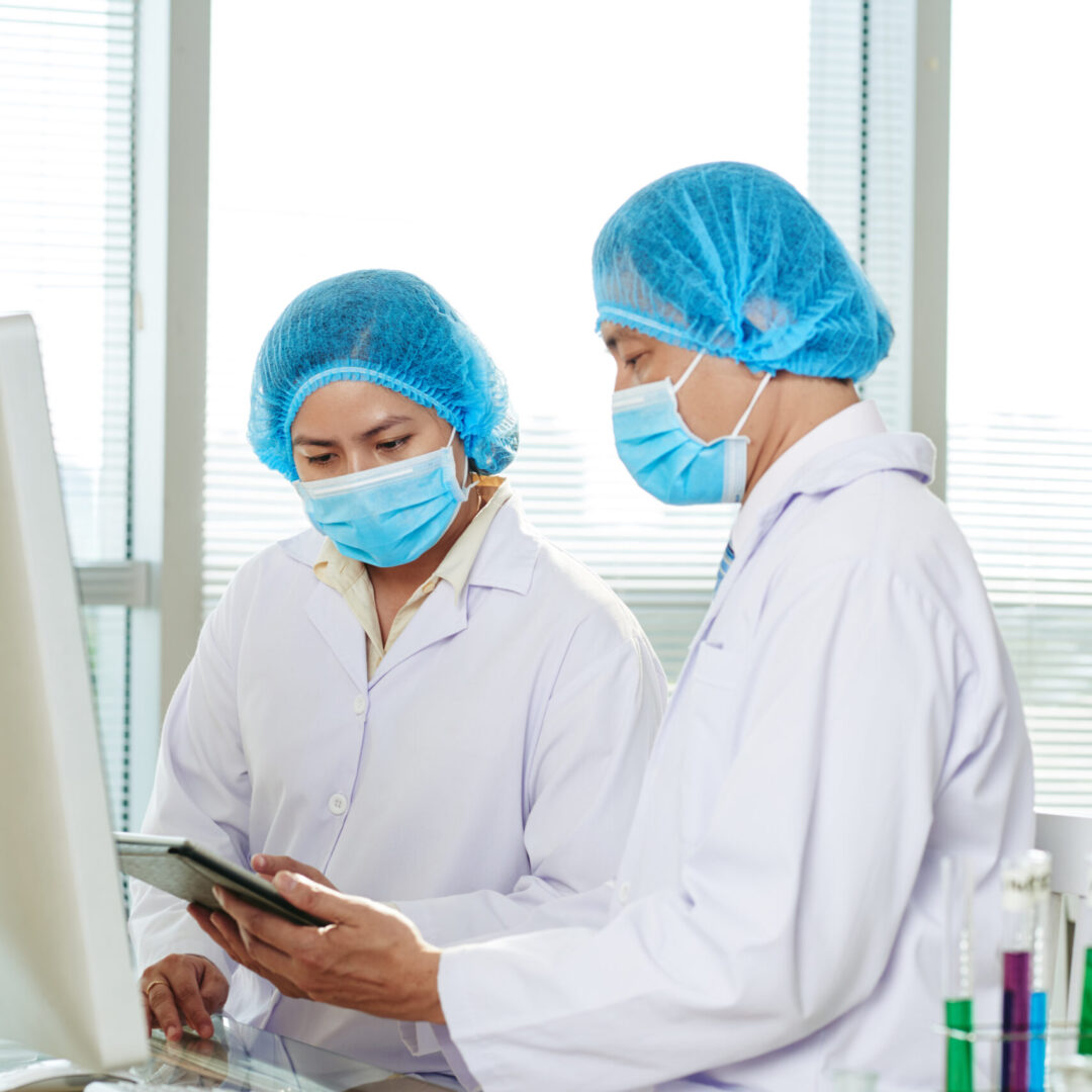 Group of hard-working researchers wearing medical masks and white coats sitting at laboratory bench and studying results of conducted experiment with help of digital tablet